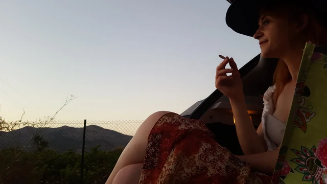 A seated person in a wide-brimmed hat beside open hills in evening light.