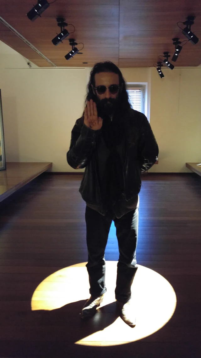 A dark-clothed person standing in a circle of light inside a gallery.