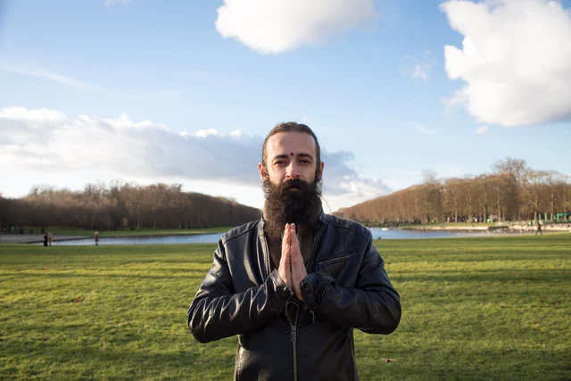 A bearded person with joined palms in an open field beneath a blue sky.
