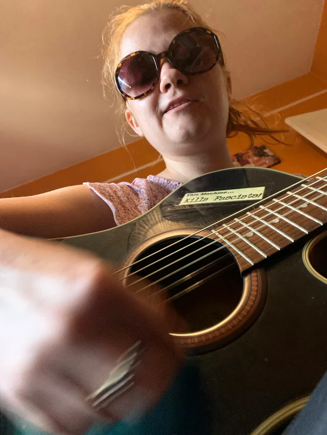 A person wearing sunglasses playing an acoustic guitar indoors.