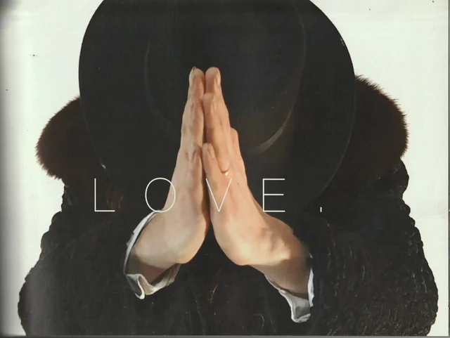 A dark-hatted figure with palms joined before their face, with the word Love.