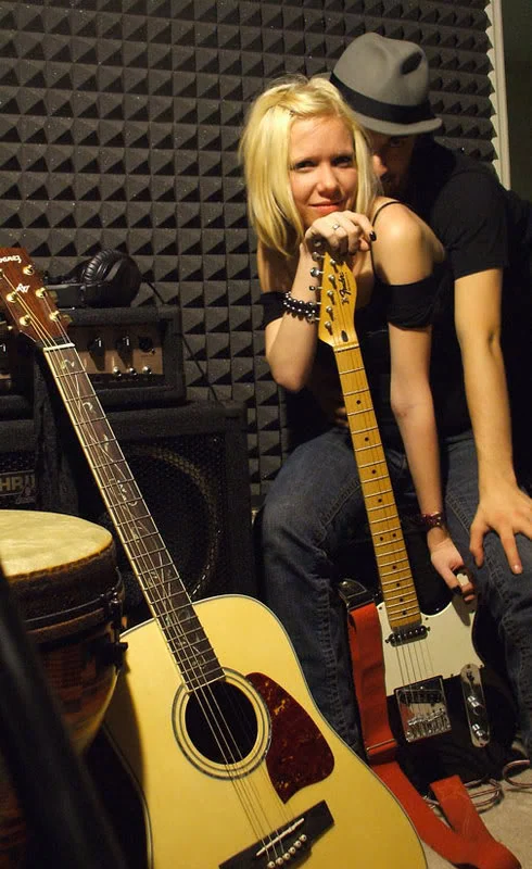 Two people posing behind acoustic and electric guitars in a music room.