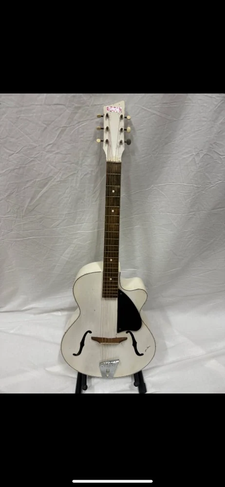 A white archtop guitar on a stand against a pale fabric backdrop.